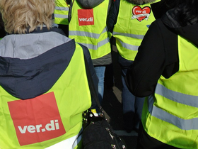 Das Bild zeigt zwei Menschen von hinten, die bei einer Streikkundgebung Warnwesten tragen. Auf einer Weste steht verdi.